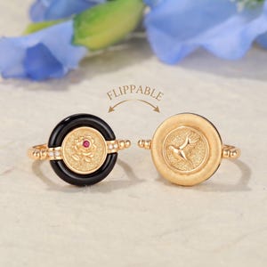May include: A flippable gold ring featuring a circular design. One side displays a black border with a central gold rose and a small red stone. The reverse side shows a solid gold textured surface with an embossed bird motif and small diamonds.