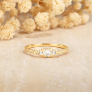 May include: A gold ring with a central, oval-shaped, light-colored stone, flanked by two smaller, round, clear stones. The ring has a delicate band and is set against a neutral background with blurred, light-colored floral elements.