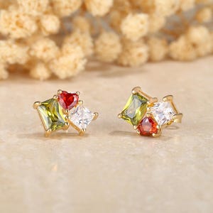 May include: A pair of gold-toned stud earrings. Each earring features a cluster of colorful gemstones: a green square, a white square, and a red heart-shaped stone. The earrings are set against a neutral background.