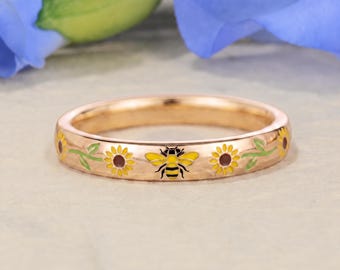Puede incluir: Un anillo de banda en oro rosa con un patrón de esmalte repetitivo de girasoles amarillos y marrones con tallos verdes y una abeja amarilla y negra en el centro. El metal tiene un acabado pulido y el diseño rodea toda la circunferencia.