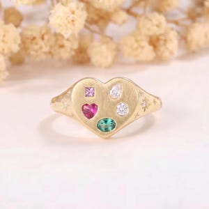 Può includere: Un anello a forma di cuore color oro con finitura spazzolata. L'anello presenta una pietra rosa quadrata, una pietra bianca a forma di pera, una pietra bianca rotonda, una pietra rosa a forma di cuore e una pietra verde ovale. Piccole stelle decorano la fascia.
