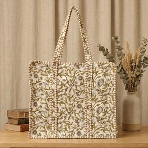 Pode incluir: Bolsa tote creme com um padrão floral em verde oliva e cinza. A bolsa tem duas alças e está sobre uma mesa de madeira. O fundo inclui um vaso com plantas secas e uma pilha de livros.