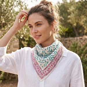 Puede incluir: Una bufanda blanca con un borde rosa y turquesa se lleva alrededor del cuello. La bufanda tiene un patrón geométrico. La persona lleva una camisa blanca abotonada. El fondo es un entorno natural al aire libre con árboles y muros de piedra.