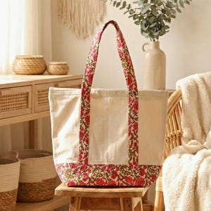 Puede incluir: Un bolso tote de color crema con un estampado floral en rojo y verde. El bolso tiene forma rectangular con cierre de cremallera y asas resistentes. Está sobre un taburete de madera.