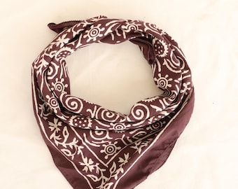 Könnte beinhalten: Ein dunkelweinrotes Bandana mit einem weißen, komplizierten Blumen- und Wirbelmuster. Das quadratische Tuch ist gefaltet und gebunden, wodurch das detaillierte Design zur Geltung kommt. Der Stoff wirkt weich und leicht, geeignet für verschiedene Zwecke.