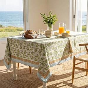 Puede incluir: Una mesa de comedor preparada para una comida, con un mantel estampado en verde y blanco, una silla de madera y una cesta de pan. Una jarra de zumo de naranja y vasos están sobre la mesa. La escena está ambientada cerca de una ventana con vistas al mar.
