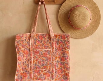 Op de afbeelding: Een totebag met bloemenprint en een strooien zonnehoed met een roze lint worden gepresenteerd tegen een neutrale muur. De totebag heeft een rechthoekige vorm met een bijpassend bloemenpatroon en een lichtroze rand. De hoed is rond.