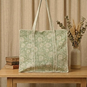 Pu&ograve; includere: Una borsa tote verde chiaro con un motivo floreale e bordi bianchi. La borsa ha manici lunghi ed &egrave; appoggiata su un tavolo di legno. Un vaso con piante secche &egrave; a destra della borsa.