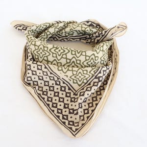 Op de afbeelding: Een gevouwen vierkante bandana met een herhalend geometrisch patroon in olijfgroen en zwart op een crèmekleurige achtergrond. De bandana is bovenaan geknoopt en toont het ontwerp. De stof lijkt zacht en licht.