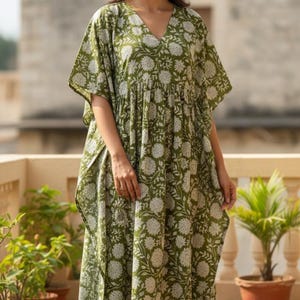 May include: A green caftan dress with a white floral pattern. The dress has a V-neck and short sleeves. The model is standing on a balcony with potted plants.