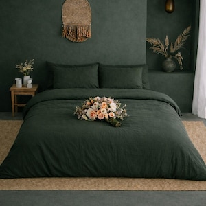 Puede incluir: Un juego de cama verde oscuro con dos almohadas y una funda nórdica, adornado con un ramo de flores. La habitación cuenta con una pared verde oscuro, un tapiz tejido y una pequeña mesita de noche de madera. Una alfombra neutra está debajo de la cama.