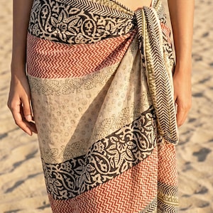 May include: A patterned sarong in shades of beige, black, red, and olive green. The fabric is wrapped and tied at the waist. The sarong features geometric and floral designs, perfect for beachwear or resort wear.