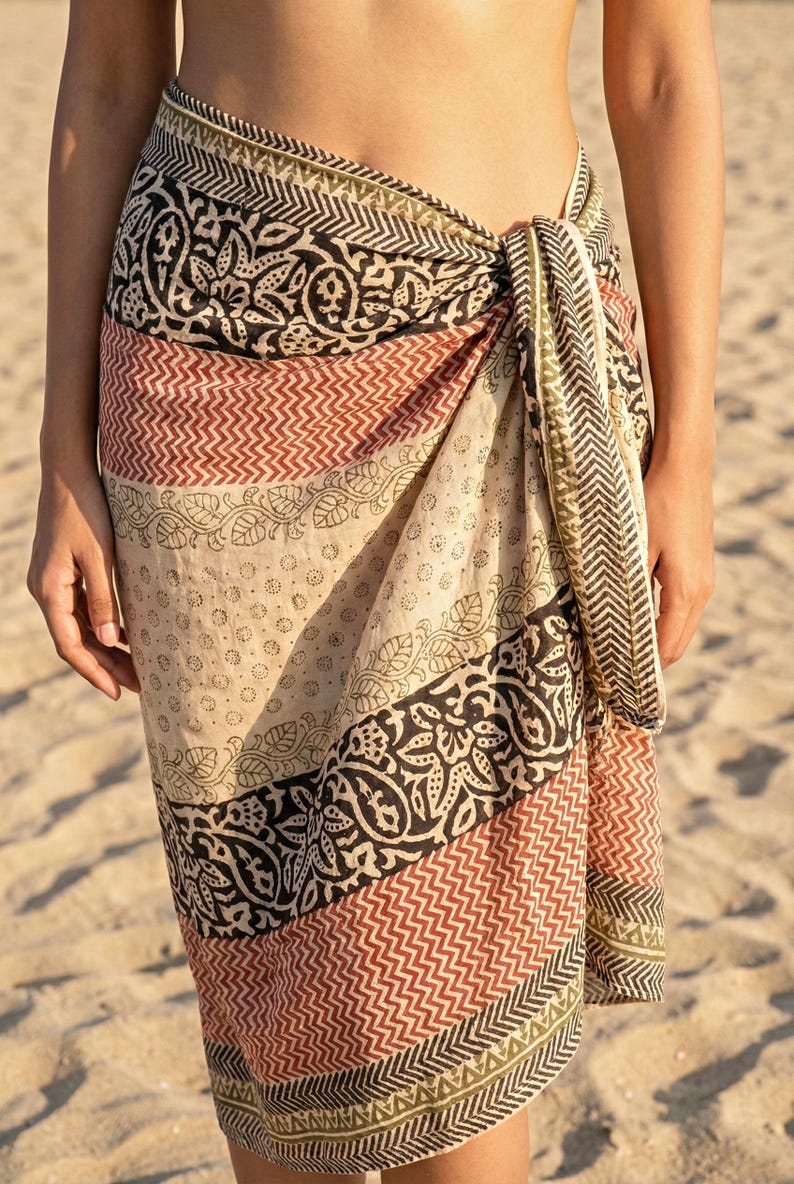 May include: A patterned sarong in shades of beige, black, red, and olive green. The fabric is wrapped and tied at the waist. The sarong features geometric and floral designs, perfect for beachwear or resort wear.