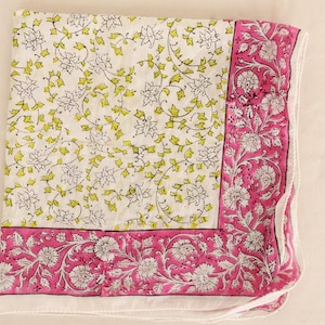 May include: A folded white textile with a floral pattern. The center features a delicate design of white flowers and yellow-green leaves. A vibrant pink border with white floral motifs frames the edges, creating a striking contrast.