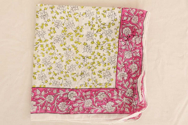 May include: A folded white textile with a floral pattern. The center features a delicate design of white flowers and yellow-green leaves. A vibrant pink border with white floral motifs frames the edges, creating a striking contrast.
