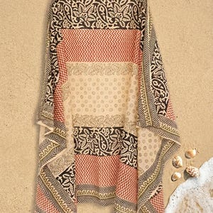 May include: A rectangular, patterned scarf with a beige background. The scarf features alternating stripes of red, black, and beige with intricate floral and geometric designs. The scarf is draped on a sandy surface.