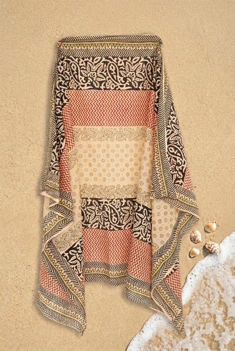 May include: A rectangular, patterned scarf with a beige background. The scarf features alternating stripes of red, black, and beige with intricate floral and geometric designs. The scarf is draped on a sandy surface.