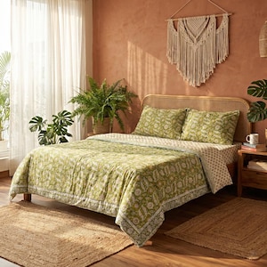 Op de afbeelding: Een bed met een groen bloemenpatroon quilt en bijpassende kussenslopen. Het bed heeft een geweven hoofdbord en staat op een houten frame. Een jute vloerkleed ligt aan het voeteneinde van het bed en een macramé wandkleed hangt boven het hoofdbord.