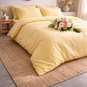 May include: A bed with a yellow duvet cover and matching pillows. A bouquet of white and pink flowers rests on the bed. A woven brown rug is on the floor. A small wooden table with mugs and a vase with flowers is on the left.