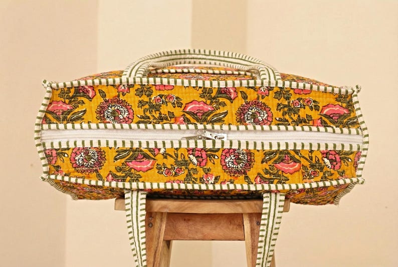 Op de afbeelding: Een gele tote bag met bloemenpatroon, een witte rits en groen-witte gestreepte rand. De tas heeft twee handvatten en een schouderriem. De tas staat op een houten kruk.