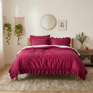 May include: A magenta bedding set featuring a duvet cover and two rectangular pillow shams, all adorned with ruffled edges. The set is displayed on a bed with a white flat sheet visible underneath the magenta duvet cover, creating a layered appearance.