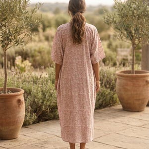 May include: A light pink, floral-patterned dress with short sleeves. The dress is worn by a person standing on a stone patio, with olive trees in terracotta pots on either side. The person has long, wavy brown hair.