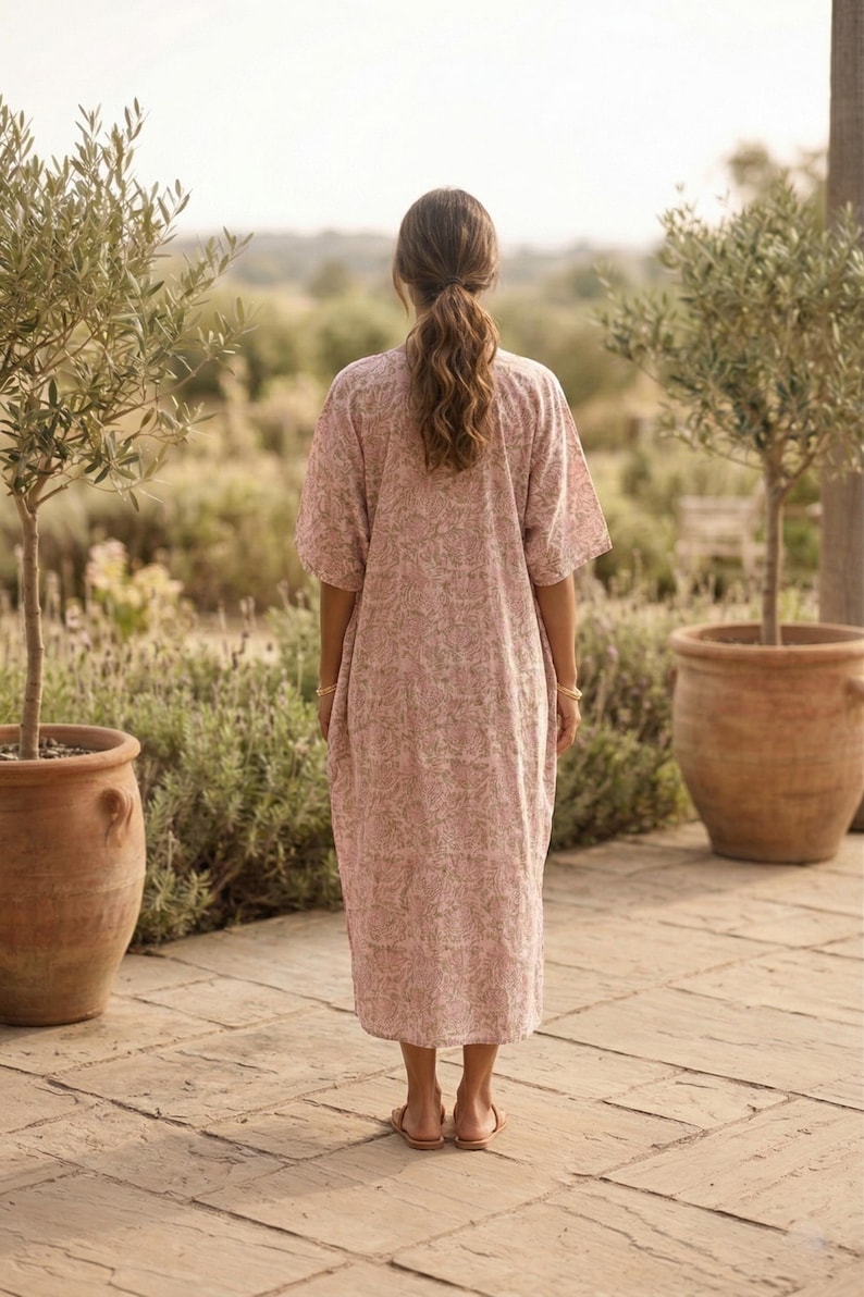 May include: A light pink, floral-patterned dress with short sleeves. The dress is worn by a person standing on a stone patio, with olive trees in terracotta pots on either side. The person has long, wavy brown hair.
