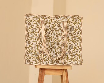 May include: A square tote bag with a floral pattern in green and brown on an off-white background. The bag has two long handles and is sitting on a small wooden stool.
