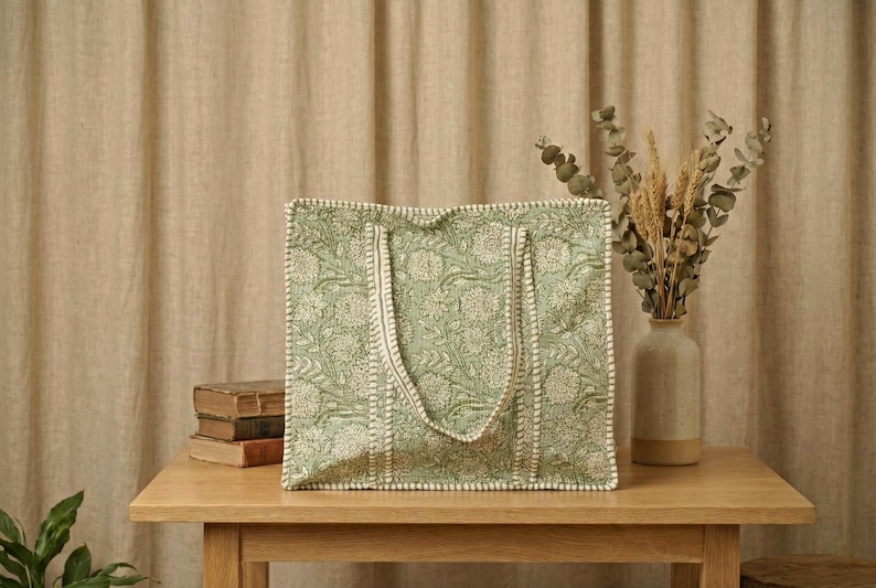 Pu&ograve; includere: Una borsa tote verde con un motivo floreale e finiture bianche &egrave; appoggiata su un tavolo di legno. La borsa ha due manici ed &egrave; accanto a una pila di libri e un vaso con piante essiccate.