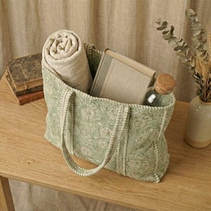 Pu&ograve; includere: Una borsa tote verde fantasia con una coperta arrotolata, un libro e una bottiglia di vetro con tappo in legno. La borsa &egrave; su un tavolo di legno, con un vaso di fiori secchi.