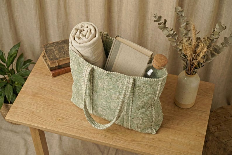 Pu&ograve; includere: Una borsa tote verde fantasia con una coperta arrotolata, un libro e una bottiglia di vetro con tappo in legno. La borsa &egrave; su un tavolo di legno, con un vaso di fiori secchi.