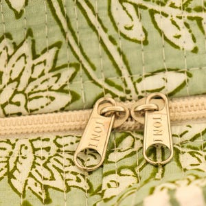 Pu&ograve; includere: Primo piano di un tessuto a fantasia floreale verde chiaro e bianco con una cerniera color crema. I tiranti della cerniera sono dorati e recano incisa la parola "TONI". Il tessuto sembra trapuntato.