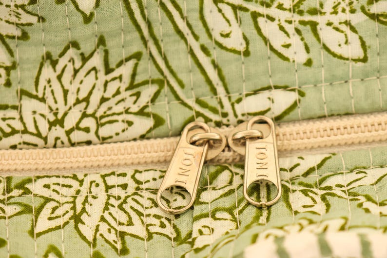 Pu&ograve; includere: Primo piano di un tessuto a fantasia floreale verde chiaro e bianco con una cerniera color crema. I tiranti della cerniera sono dorati e recano incisa la parola "TONI". Il tessuto sembra trapuntato.