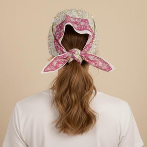May include: A floral print headscarf with a pink and white paisley design tied in a bow. The headscarf is worn over long, wavy brown hair. The background is a solid beige color.