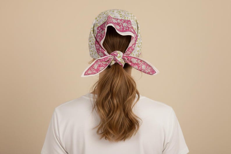 May include: A floral print headscarf with a pink and white paisley design tied in a bow. The headscarf is worn over long, wavy brown hair. The background is a solid beige color.