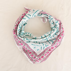 Op de afbeelding: Witte bandana met een roze bloemenrand en turquoise abstracte vormen. De vierkante sjaal is opgevouwen en geknoopt, waarbij het patroon wordt getoond. Dit accessoire kan om de nek of op het hoofd worden gedragen.
