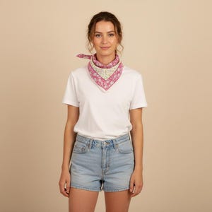 May include: A woman wearing a white t-shirt, light blue denim shorts, and a pink and white floral patterned scarf tied around her neck. The scarf is square-shaped and adds a pop of color to the outfit.