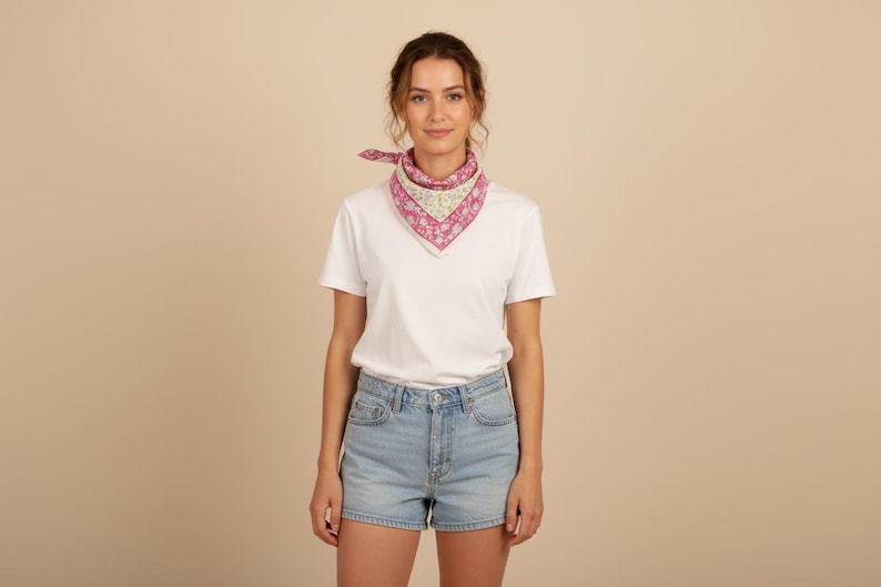 May include: A woman wearing a white t-shirt, light blue denim shorts, and a pink and white floral patterned scarf tied around her neck. The scarf is square-shaped and adds a pop of color to the outfit.