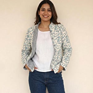 Könnte beinhalten: Eine Frau trägt ein weißes Oberteil und eine dunkelblaue Jeans. Sie trägt eine cremefarbene Jacke mit blauem Blumenmuster. Das Innenfutter der Jacke hat ein Streifenmuster.