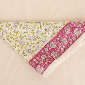 May include: Folded white and pink floral patterned fabric. The fabric features a white background with yellow and green floral designs on one side and a pink border with white floral designs on the other. The fabric is folded into a triangle.