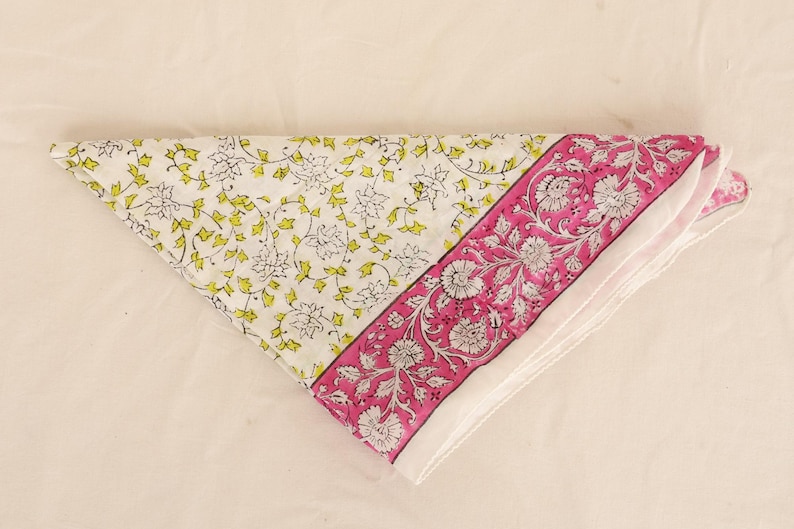 May include: Folded white and pink floral patterned fabric. The fabric features a white background with yellow and green floral designs on one side and a pink border with white floral designs on the other. The fabric is folded into a triangle.