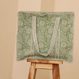 Pu&ograve; includere: Una borsa tote verde con un motivo floreale e cinghie bianche. La borsa &egrave; in tessuto e ha una forma rettangolare. La borsa &egrave; appoggiata su uno sgabello di legno.