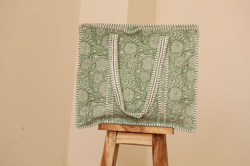 Pu&ograve; includere: Una borsa tote verde con un motivo floreale e cinghie bianche. La borsa &egrave; in tessuto e ha una forma rettangolare. La borsa &egrave; appoggiata su uno sgabello di legno.