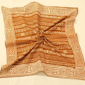 Op de afbeelding: Een vierkante sjaal met een bruin en wit patroon. De sjaal heeft een herhalend geometrisch en tribaal ontwerp. De rand heeft een vierkant en bloemenpatroon. De sjaal is gevouwen en gedrapeerd op een lichtbeige oppervlak.