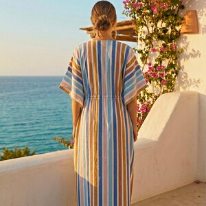 May include: A long, striped dress in shades of blue, tan, and pink. The dress has a drawstring waist and short sleeves. The person is standing on a white balcony overlooking the ocean. Bougainvillea flowers bloom on the wall.