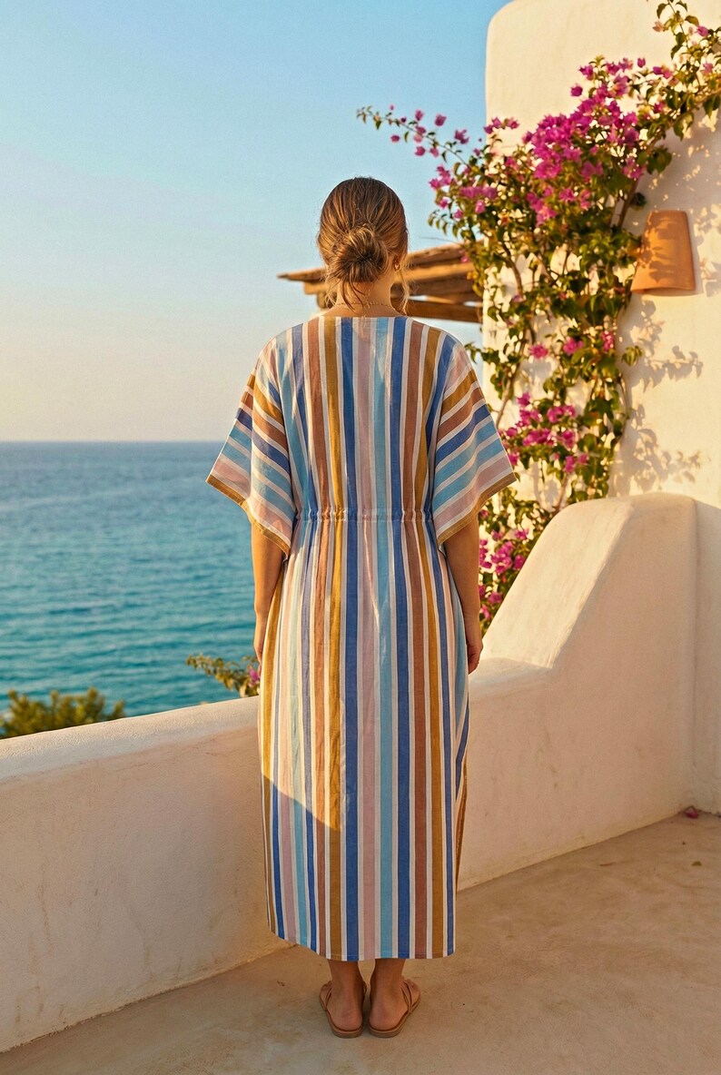 May include: A long, striped dress in shades of blue, tan, and pink. The dress has a drawstring waist and short sleeves. The person is standing on a white balcony overlooking the ocean. Bougainvillea flowers bloom on the wall.