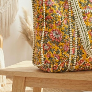 Op de afbeelding: Een grote tote bag met een bloemenpatroon in tinten geel, roze en groen. De tas heeft een gewatteerd ontwerp en gestreepte handvatten. De tas staat op een houten tafel.