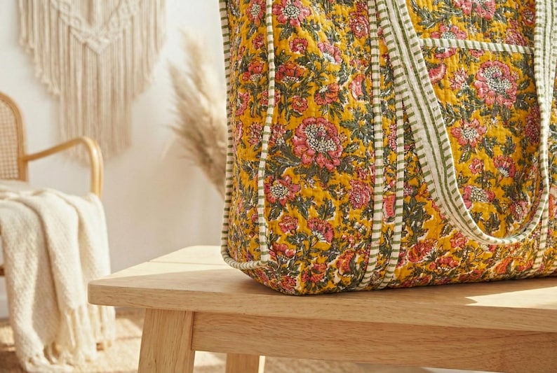 Op de afbeelding: Een grote tote bag met een bloemenpatroon in tinten geel, roze en groen. De tas heeft een gewatteerd ontwerp en gestreepte handvatten. De tas staat op een houten tafel.