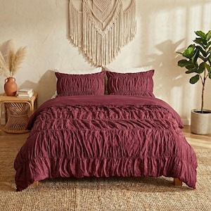 May include: A burgundy bedspread and two pillowcases with a ruffled design. The bedding is on a wooden bed frame, with a woven rug underneath. A macrame wall hanging is above the bed, and a potted plant is to the right.