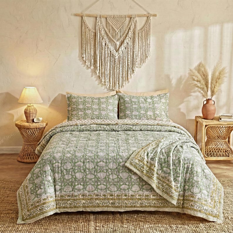 May include: A bedroom scene featuring a green and white floral patterned quilt set with matching pillowcases. A macrame wall hanging and a woven side table with a lamp add to the bohemian aesthetic. The bedspread has a decorative border.